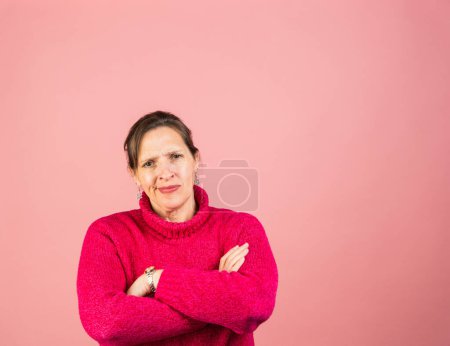 A mature woman in her early fifties with a pink turtleneck looking at the camera exasperated with hands folded, tucking in her chin to expose the double chin