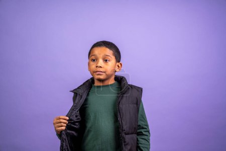 Téléchargez les photos : Un jeune garçon portant une chemise verte et un gilet noir se tient devant un fond violet. Il semble sérieux et concentré - en image libre de droit