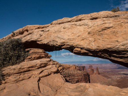 Mesa Arch in the desert with a view of the mountains. The archway is made of rocks and the sky is blue