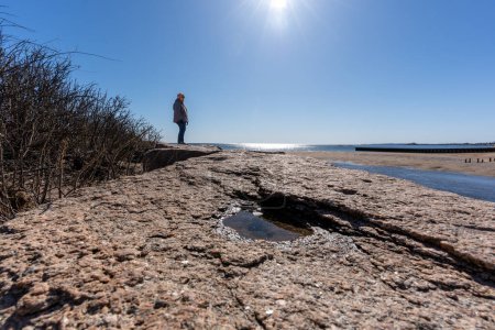 Téléchargez les photos : Une personne est debout sur une plage rocheuse, regardant l'océan. Le ciel est clair et bleu, et le soleil brille - en image libre de droit
