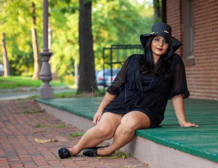 Téléchargez les photos : Une femme asiatique du sud plus la taille s'assoit sur une plate-forme de train regardant la caméra portant une robe noire courte. - en image libre de droit