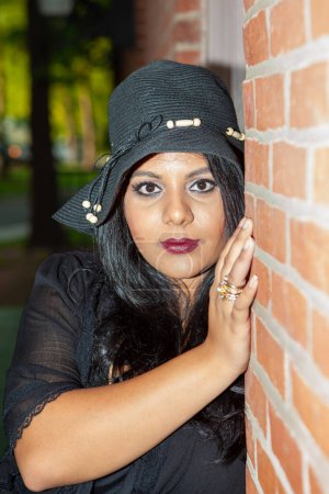 Téléchargez les photos : Une Indienne d'Asie du Sud en robe noire porte un chapeau noir et des anneaux d'or. Elle s'appuie contre un mur de briques - en image libre de droit
