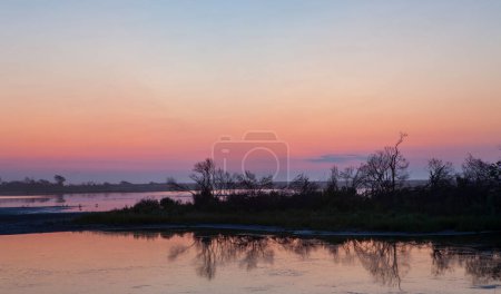 Téléchargez les photos : Un beau coucher de soleil sur un lac avec des arbres en arrière-plan. Le ciel est un mélange de teintes rose et orange, créant une atmosphère sereine et paisible. L'eau est calme, reflétant les couleurs du ciel - en image libre de droit