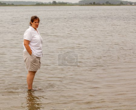 Téléchargez les photos : Une femme se tient dans l'eau, regardant vers l'horizon. L'eau est calme et le ciel couvert - en image libre de droit