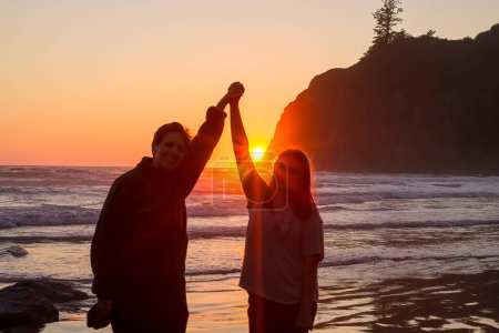Téléchargez les photos : Mère et fille debout sur la plage, tenant la main et souriant au coucher du soleil. Scène est heureuse et romantique - en image libre de droit