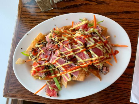 Téléchargez les photos : Une assiette de nachos au thon Ahi avec une sauce jaune et des carottes sur le dessus. La plaque est blanche et la nourriture est sur une table en bois - en image libre de droit