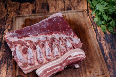 Téléchargez les photos : Ventre de porc frais sur une planche à découper usagée reposant sur une vieille table en bois - en image libre de droit