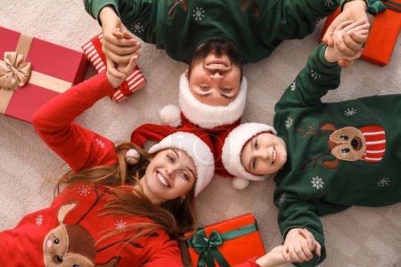 Téléchargez les photos : Joyeux parents avec leur petit fils dans les chapeaux de Père Noël tenant la main à la maison la veille de Noël, vue sur le dessus - en image libre de droit