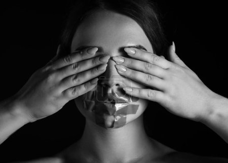 Young woman with taped mouth covering her eyes on dark background. Censorship concept