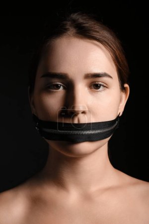 Junge Frau mit Reißverschluss am Mund vor dunklem Hintergrund. Zensurkonzept