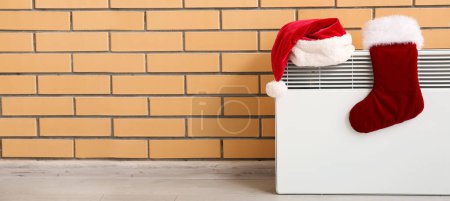 Photo for Santa hat and Christmas sock on electric radiator near brick wall with space for text - Royalty Free Image