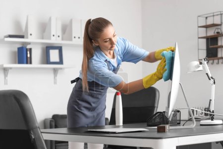 Téléchargez les photos : Travailleuse du service de nettoyage essuyant l'écran d'ordinateur dans le bureau - en image libre de droit