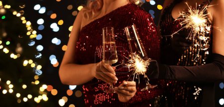 Téléchargez les photos : Femmes dans des robes élégantes, avec des étincelles brûlantes et des verres de champagne célébrant Noël ensemble - en image libre de droit
