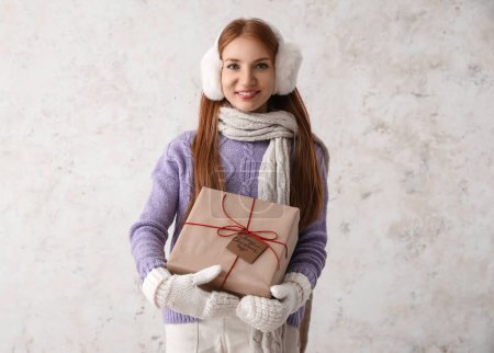 Foto de Mujer pelirroja joven en orejeras calientes con regalo de Navidad sobre fondo claro - Imagen libre de derechos
