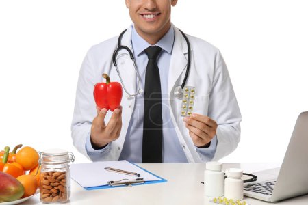Photo for Male doctor with vegetables and vitamins sitting at table on white background - Royalty Free Image