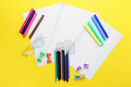Téléchargez les photos : Feuilles de papier vierges avec lunettes, crayons, feutres, gommes et aiguiseurs sur fond jaune - en image libre de droit