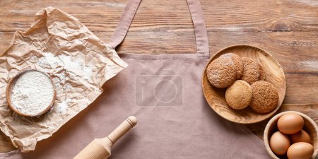 Foto de Delantal limpio, rodillo, huevos, harina y galletas sobre fondo de madera, vista superior - Imagen libre de derechos