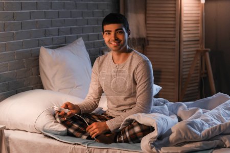 Junger Mann sitzt nachts auf elektrischem Heizkissen im Schlafzimmer