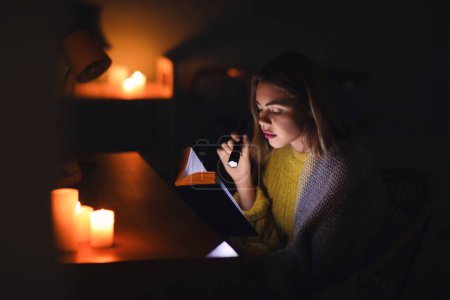 Téléchargez les photos : Jeune femme lecture livre avec lampe de poche à la maison pendant la panne d'électricité - en image libre de droit