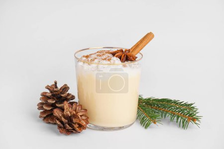 Glass of tasty eggnog cocktail, fir branch and cones on white background