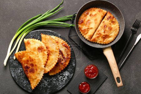 Frying pan and board with tasty chebureks, sauces and onion on dark grunge background