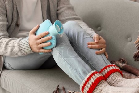 Young man warming his knee with hot water bottle on sofa at home, closeup