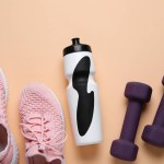 Sports water bottle with dumbbells and sneakers on beige background