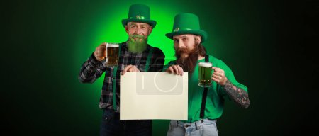 Foto de Bearded men with blank poster and glasses of beer on dark green background. St. Patrick's Day celebration - Imagen libre de derechos