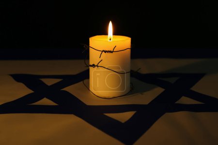 Burning candle with barbed wire on flag of Israel against dark background. International Holocaust Remembrance Day