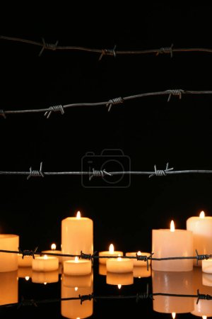 Barbed wire and burning candles on glass table against dark background. International Holocaust Remembrance Day