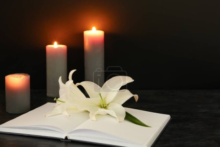 Aufgeschlagenes Buch, weiße Lilienblumen und brennende Kerzen auf dunklem Hintergrund