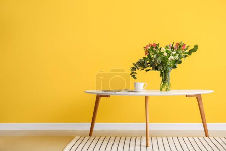 Foto de Florero con hermosas flores de alstroemeria, taza de café y revista en la mesa cerca de la pared amarilla - Imagen libre de derechos