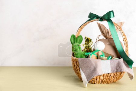 Foto de Cesta con conejito, huevos de Pascua, caramelos y bolsa de regalo sobre mesa verde contra pared beige - Imagen libre de derechos