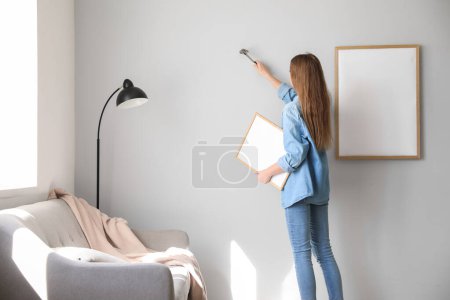 Téléchargez les photos : Jeune femme avec cadre vierge clouant sur le mur gris à la maison - en image libre de droit