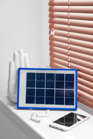 Portable solar panel charging mobile phone and earphones on windowsill in room