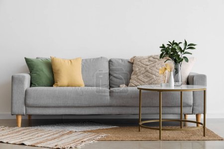 Téléchargez les photos : Canapé confortable gris avec coussins et branches de plantes dans un vase sur la table près du mur clair - en image libre de droit