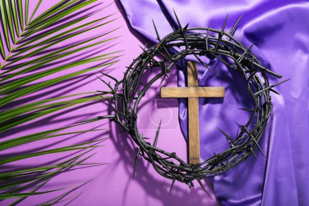 Foto de Cruz de madera con corona de espinas, hoja de palma y tela púrpura sobre fondo violeta. Concepto Viernes Santo - Imagen libre de derechos