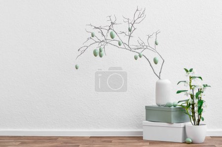 Vase with tree branch, Easter eggs and houseplant near light wall