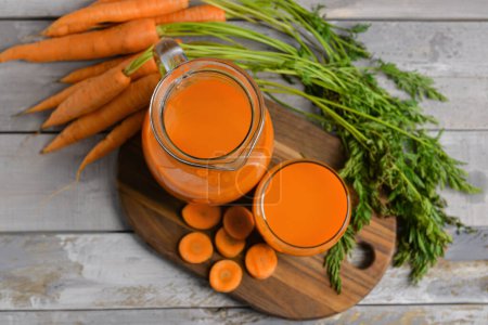 Glass and jug of fresh carrot juice on grey wooden background
