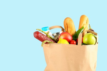 Paper bag with vegetables, fruits, sausage, bread and milk on light blue background