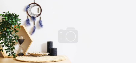 Photo for Stylish dream catcher hanging on white wall and table with shelf, candles and hourglass. Banner for design - Royalty Free Image