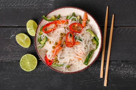 Photo for Bowl with tasty rice noodles, chopsticks, vegetables and lime on black wooden table - Royalty Free Image