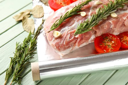 Aluminium foil roll with piece of raw meat, tomatoes, rosemary and spices on color wooden background, closeup