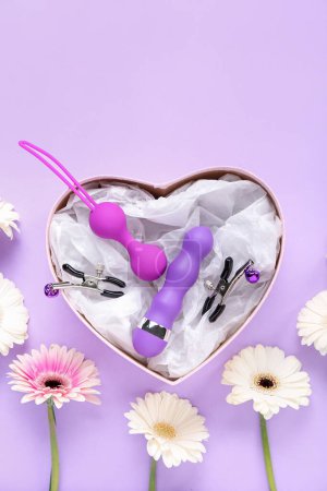 Téléchargez les photos : Coffret cadeau avec des jouets sexuels et des fleurs sur fond lilas - en image libre de droit