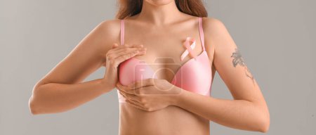 Woman in underwear and with pink ribbon checking her breast on grey background. Cancer awareness concept