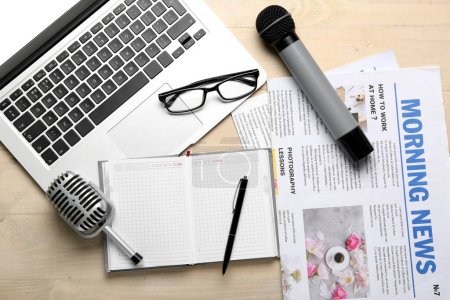 Laptop mit Brille, Mikrofonen, Notizbuch und Zeitungen auf hellem Holzhintergrund