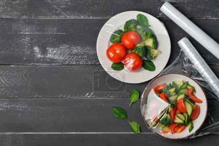 Schüssel und Teller mit Salat umwickelt mit Stretchfolie auf dunklem Holzhintergrund