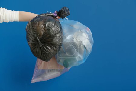 Mano femenina sosteniendo bolsas llenas de basura sobre fondo azul