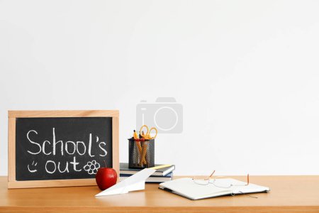 Kreidetafel mit Text SCHULE 'S OUT, Apfel, Papierflieger und Schreibwaren auf Tisch neben Lichtwand