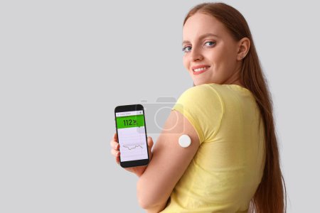 Téléchargez les photos : Femme avec capteur de glucose utilisant un téléphone portable pour mesurer le taux de sucre dans le sang sur fond gris - en image libre de droit